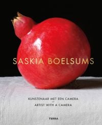 Shiny red pomegranate on white table cloth, black background, SASKIA BOELSUMS in gold font to centre, ARTIST WITH A CAMERA in black font below.