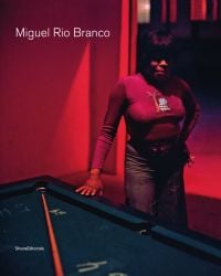 Black female with hand resting on pool table, in dark lit room, 'Miguel Rio Branco', in white font to upper left, by Silvana.