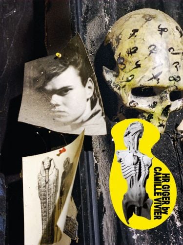 Photograph of H.R Giger pinned to wall, skull with black symbols, HR GIGER BY CAMILLE VIVIER, in black font on yellow banner to bottom right.