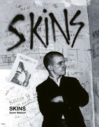 Young white skinhead wearing black jacket, arms folded, standing in front of wall graffitied with 'SKINS', in black spray paint.
