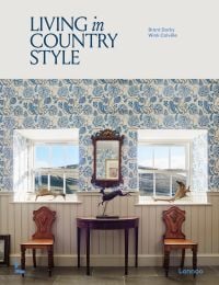 Book cover of Living in Country Style, with interior with white panelled walls, blue floral wallpaper, and two windows looking out to hilly landscape. Published by Lannoo Publishers.