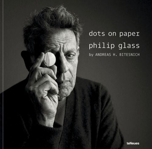 White male in dark jacket, holding circular piece of glass to right eye, on cover of 'dots on paper, Philip Glass by Andreas H. Bitesnich', by teNeues Books.