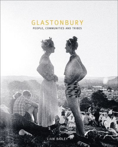 Two white female festival goers standing at top of hill, sea of people and tents behind them, on cover of 'Glastonbury', by ACC Art Books.
