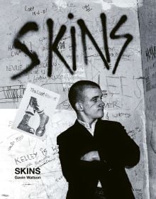 Young white male skinhead in black jacket, looking to his left, arms folded, 'SKINS', in black graffiti on wall behind, ACC Art Books.