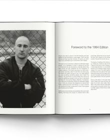 Young white male skinhead in black jacket, looking to his left, arms folded, 'SKINS', in black graffiti on wall behind, ACC Art Books.