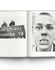 Young white male skinhead in black jacket, looking to his left, arms folded, 'SKINS', in black graffiti on wall behind, ACC Art Books.