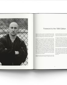 Young white male skinhead wearing black jacket, arms folded, standing in front of wall graffitied with 'SKINS', in black spray paint, by ACC Art Books.