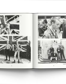 Young white male skinhead wearing black jacket, arms folded, standing in front of wall graffitied with 'SKINS', in black spray paint, by ACC Art Books.