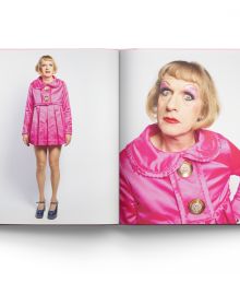 Grayson Perry wearing pink silk dress and make-up, holding baby, on cover of 'MUSE, A Portrait of Grayson Perry', by ACC Art Books.