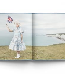 Grayson Perry wearing pink silk dress and make-up, holding baby, on cover of 'MUSE, A Portrait of Grayson Perry', by ACC Art Books.