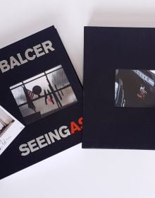 Row of knives and a machete, on magnetic strip, American flag and globe to left, on black cover of 'Seeing As, René Balcer', by ACC Art Books.