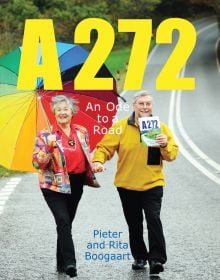 Book cover of A272-An Ode to a Road, with Pieter and Rita Boogaart walking hand-in-hand down the A272, Rita holding a large rainbow umbrella. Published by Pallas Athene.