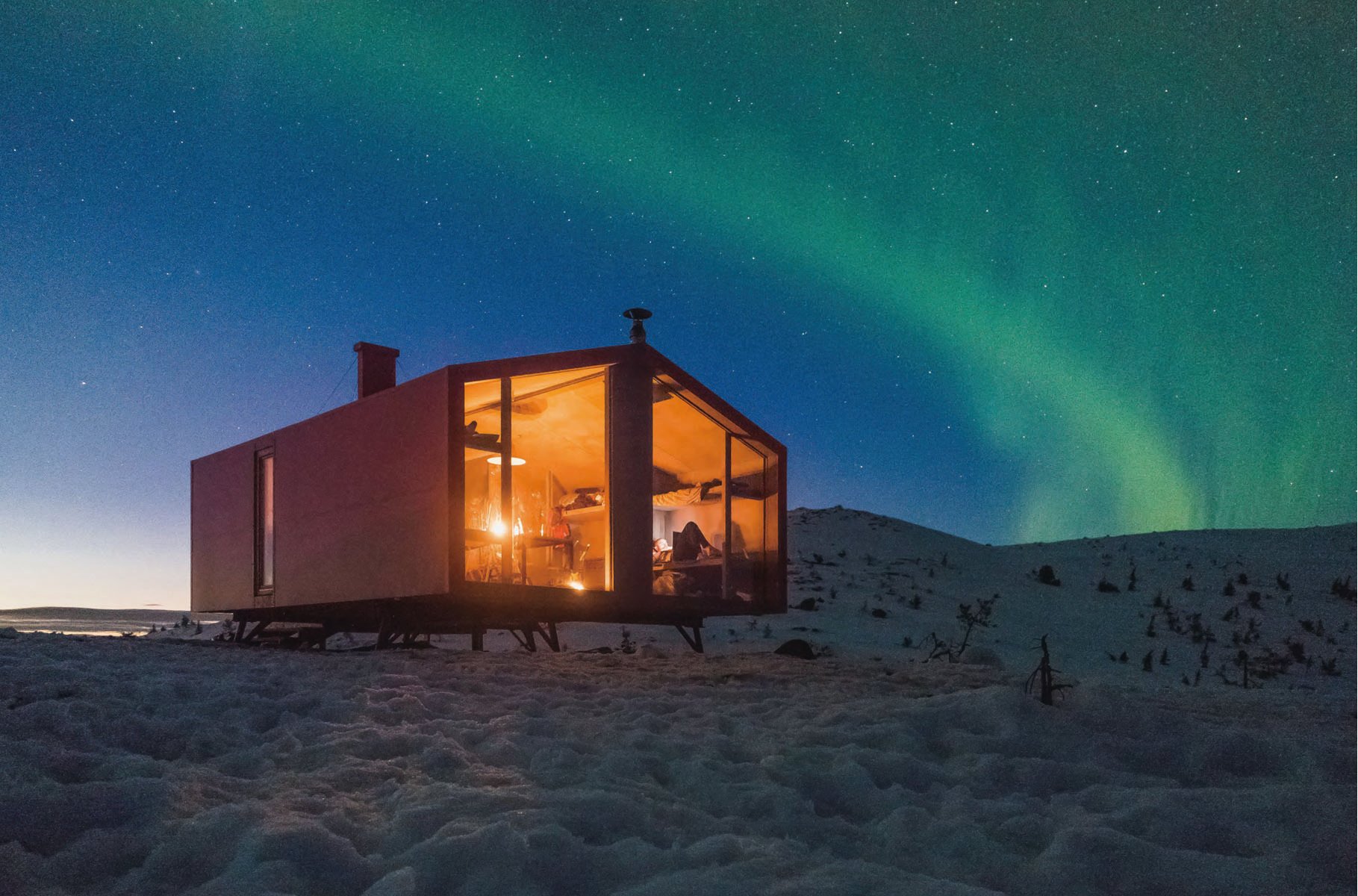 Modern cabin, with interior lights on, in forest at dusk, Cabins in white font to upper left.