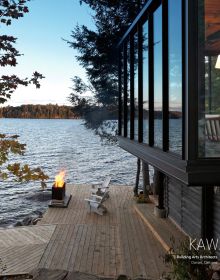 Stunning shot of modern glass building with flat moss roof poking out of Canadian forest near lake, Northern Hideaways in blue font above.