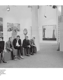 Bridget Riley sitting on a table, 'Bridget Riley: A Very Very Person', in white, and pink font to left side, by Ridinghouse.