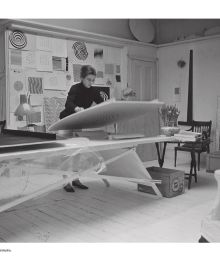 Bridget Riley sitting on a table, 'Bridget Riley: A Very Very Person', in white, and pink font to left side, by Ridinghouse.