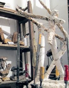 Photograph of industrial rock sculpture with rusted metal poles, in white exhibition space, 'OLGA JEVRI?', in orange font to upper left, by Ridinghouse.