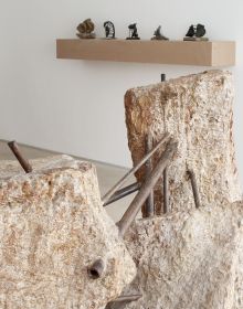 Photograph of industrial rock sculpture with rusted metal poles, in white exhibition space, 'OLGA JEVRI?', in orange font to upper left, by Ridinghouse.