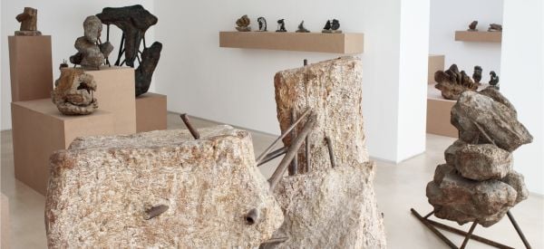 Photograph of industrial rock sculpture with rusted metal poles, in white exhibition space, 'OLGA JEVRI?', in orange font to upper left, by Ridinghouse.