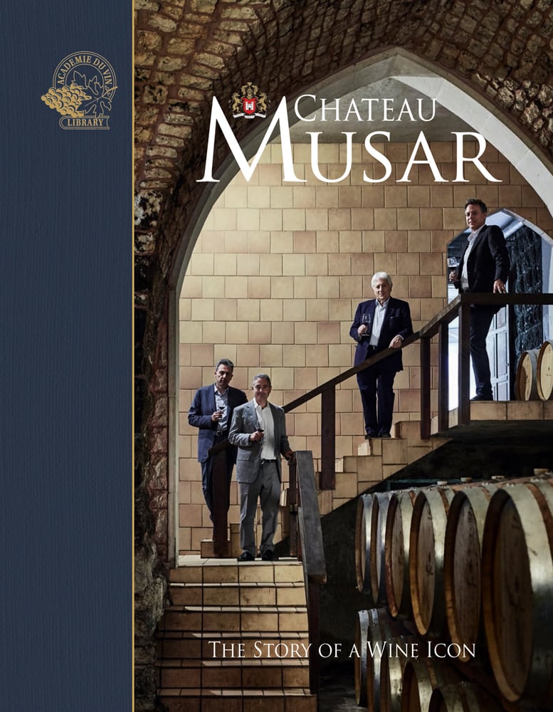 Four suited men holding glasses of wine, standing on steps of barrel store, Chateau Musar, in white font above.