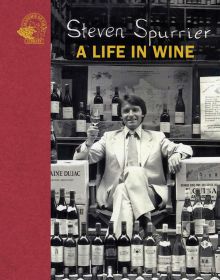 Steven Spurrier in white suit sitting in chair holding glass of wine and surrounded by stacks of wine, on cover of 'Steven Spurrier, A Life in Wine', by Academie du Vin Library.