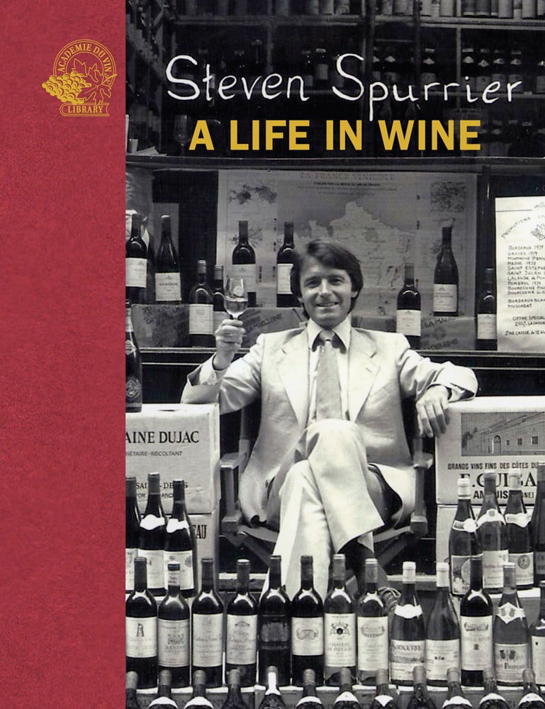 Steven Spurrier in white suit sitting in chair holding glass of wine and surrounded by stacked full wine bottles and Steven Spurrier A Life in Wine in white and yellow font above