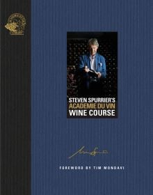 Man wearing blue jacket, standing in wine cellar, holding a bottle wine, on blue cover of 'Steven Spurrier's Académie du Vin Wine Course, The Art of Learning by Tasting', by Academie du Vin Library.