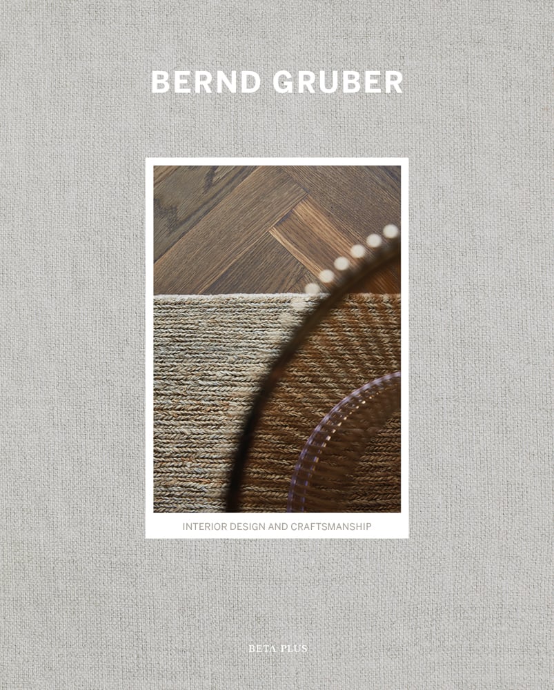 Woven beige rug on dark herringbone flooring, grey linen cover, BERND GRUBER in white font above.