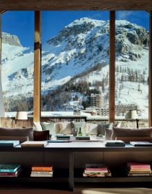 Book cover of Alpine Elegance: High-End Mountain Homes, featuring a cozy house with a view of snow topped mountains through the large windows. Published by Beta-Plus.