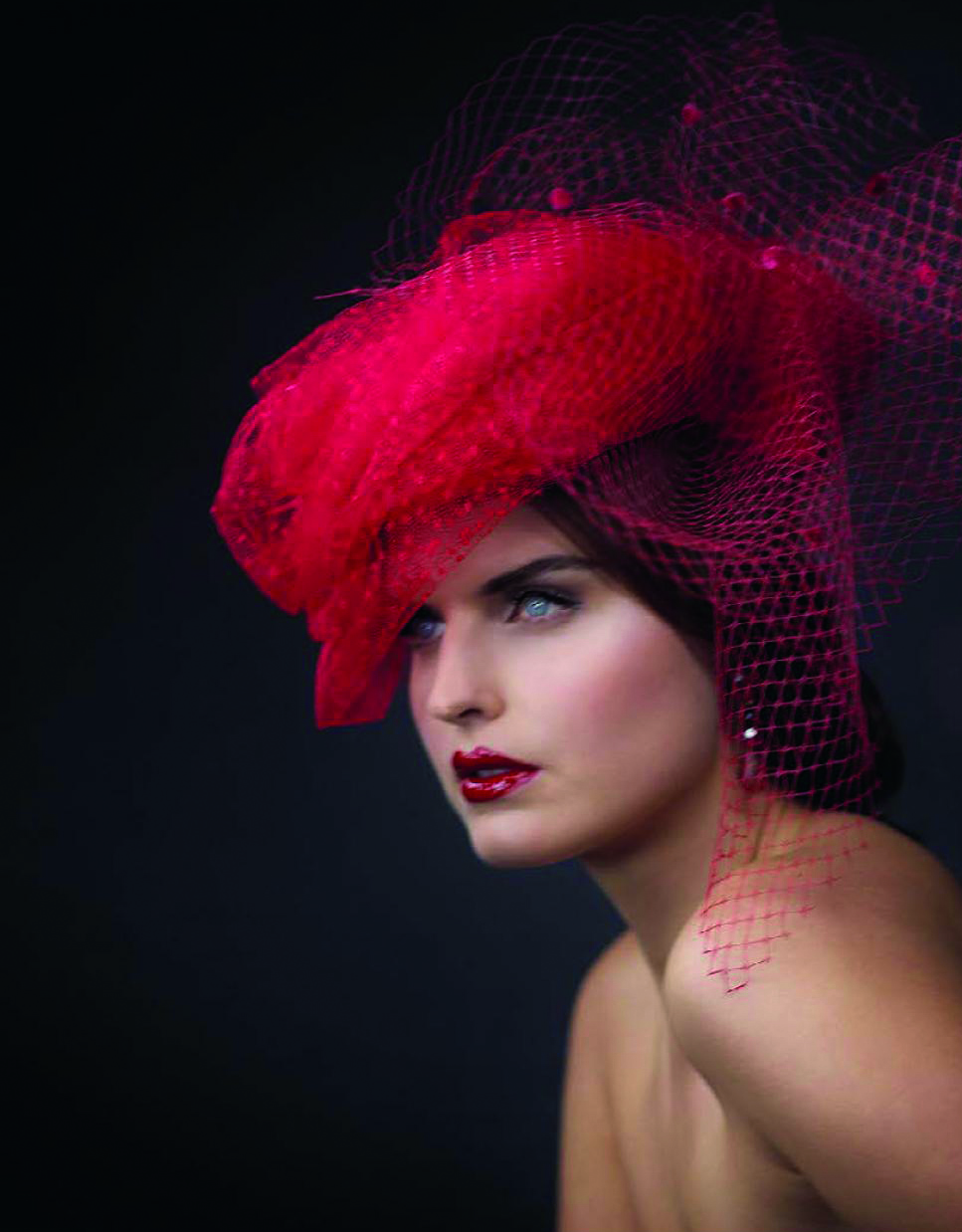 Fashion model in brown, blue and green feathered hat with black silk bow to centre, FABIENNE DELVIGNE in white font to lower right.