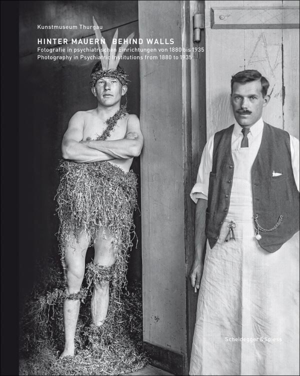 Book cover of Behind Walls: Photography in Psychiatric Institutions from 1880 to 1935, with a man in long white smock with waistcoat, and man in tribal dress, standing in doorway. Published by Scheidegger & Spiess.