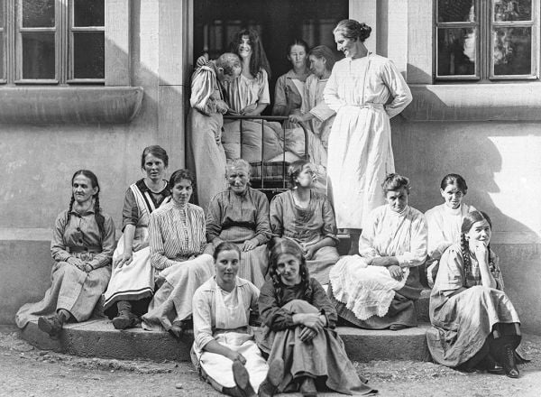 Book cover of Behind Walls: Photography in Psychiatric Institutions from 1880 to 1935, with a man in long white smock with waistcoat, and man in tribal dress, standing in doorway. Published by Scheidegger & Spiess.