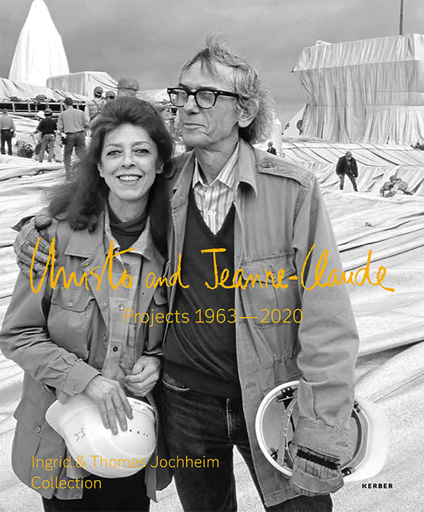 Christo putting arm around Jeanne-Claude, both holding white hard hats, installation space with workers behind, Christo and Jeanne-Claude Projects 1963-2020 in orange font to centre.
