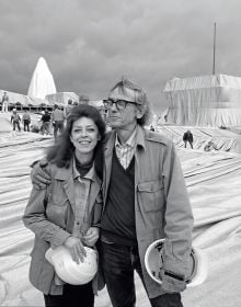Christo putting arm around Jeanne-Claude, both holding white hard hats, installation space with workers behind, Christo and Jeanne-Claude Projects 1963-2020 in orange font to centre.