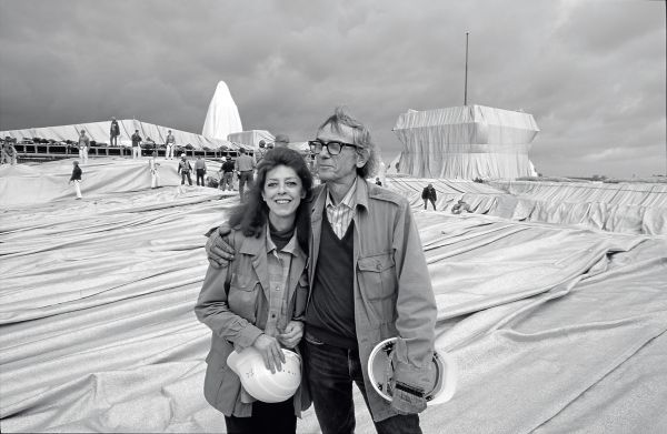 Christo putting arm around Jeanne-Claude, both holding white hard hats, installation space with workers behind, Christo and Jeanne-Claude Projects 1963-2020 in orange font to centre.