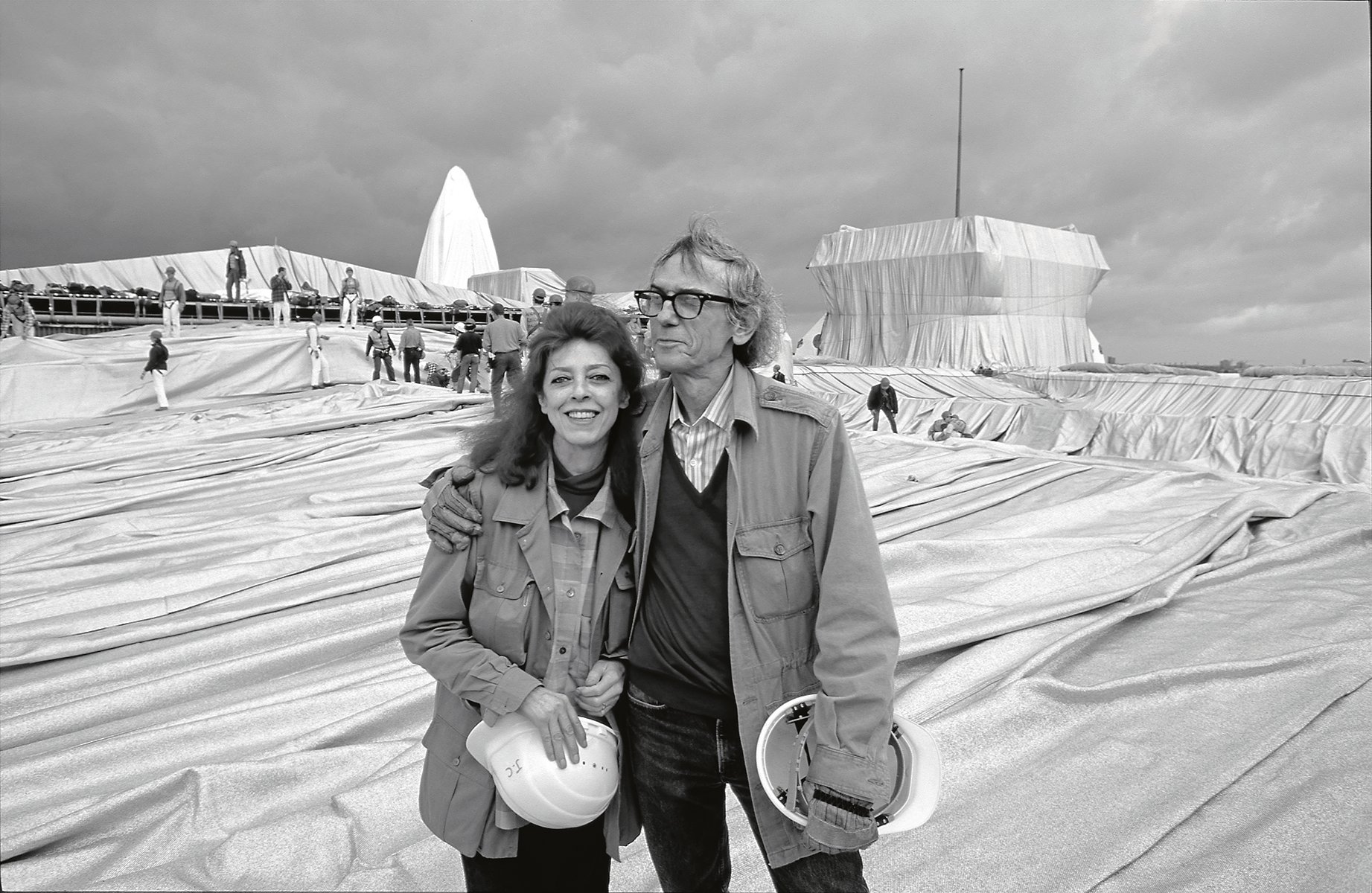 Christo putting arm around Jeanne-Claude, both holding white hard hats, installation space with workers behind, Christo and Jeanne-Claude Projects 1963-2020 in orange font to centre.