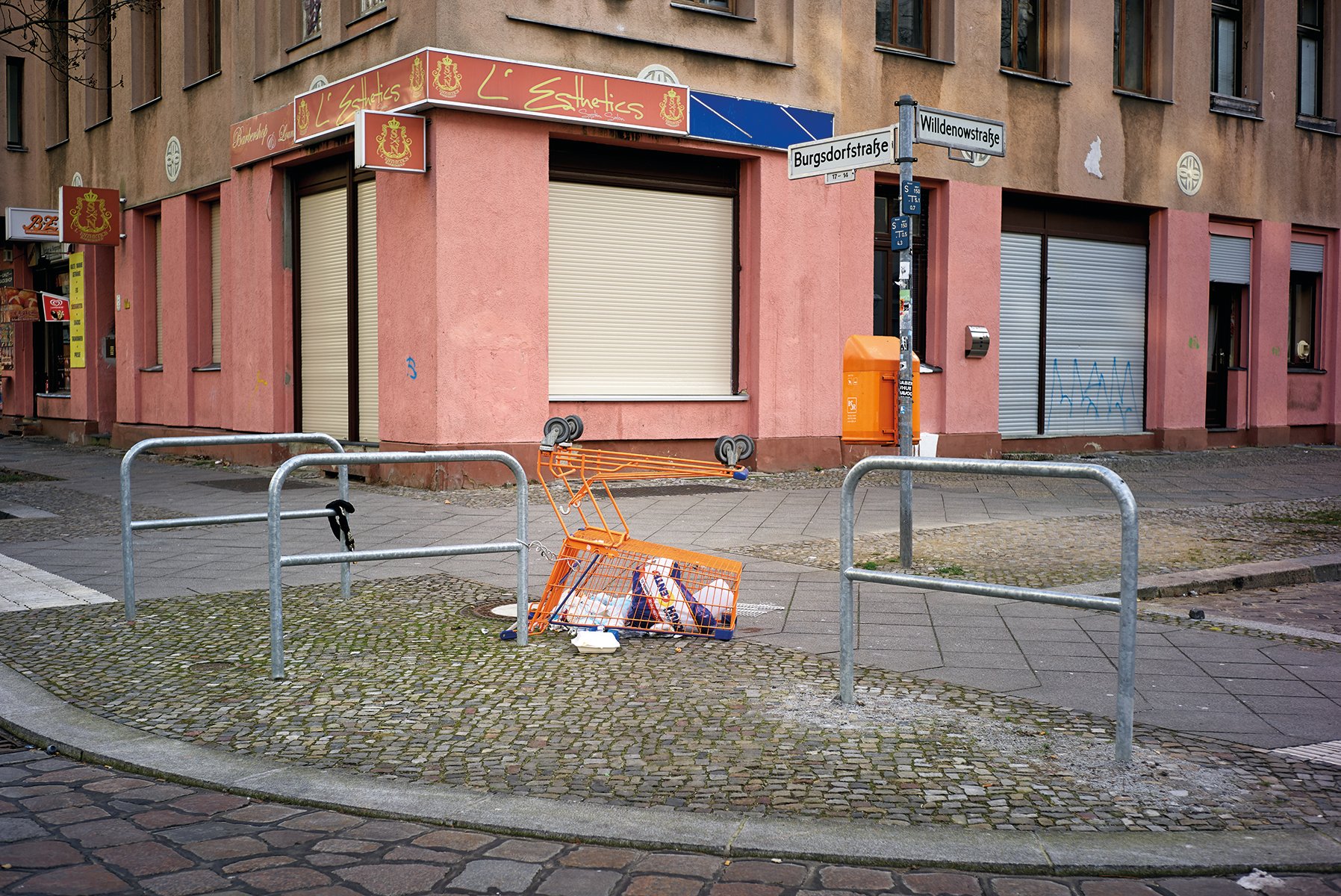 2 empty shopping trollies abandoned on corner of grey brick building with Einkaufswagen Luca Ellena in white font on coral banner to left side