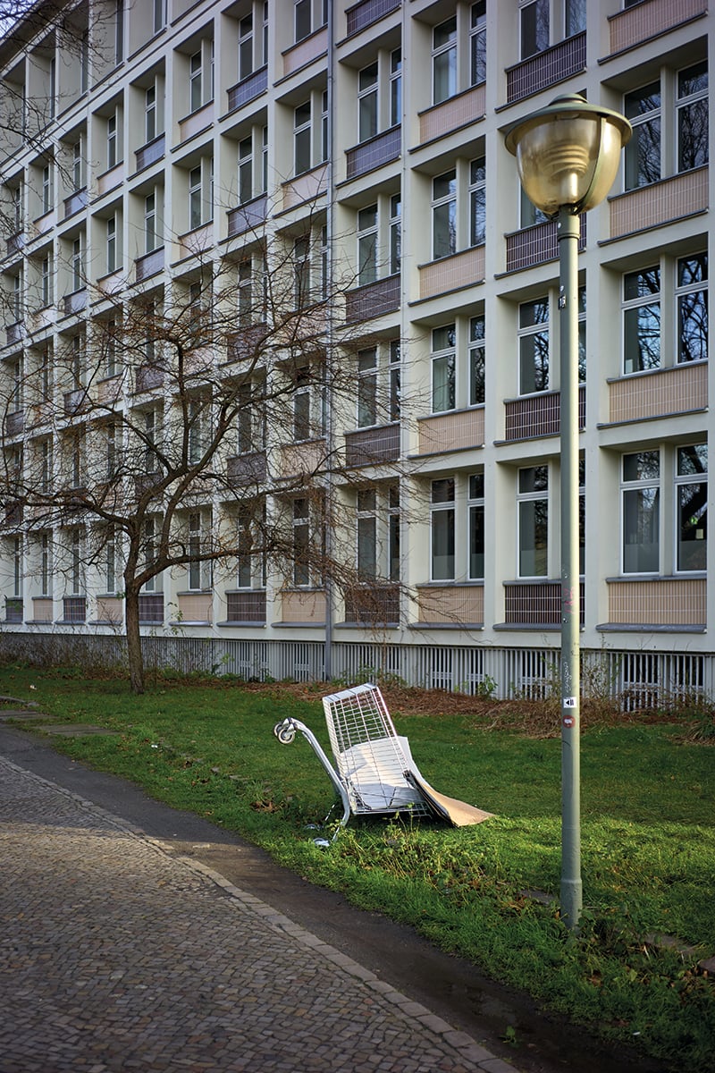 2 empty shopping trollies abandoned on corner of grey brick building with Einkaufswagen Luca Ellena in white font on coral banner to left side