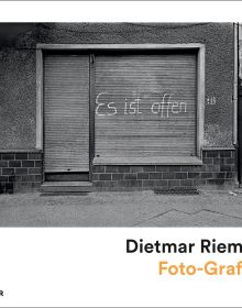 A shuttered-up shop in Ost-Berlin, 1986, 'es ist offen' spray painted on shutter door in white, on white landscape cover of 'Dietmar Riemann, Foto-Grafiker', by Kerber.