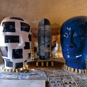 Ceramic glazed sculpture of head on low table, orange cover, Jun Kaneko The Space Between in white font to upper left.