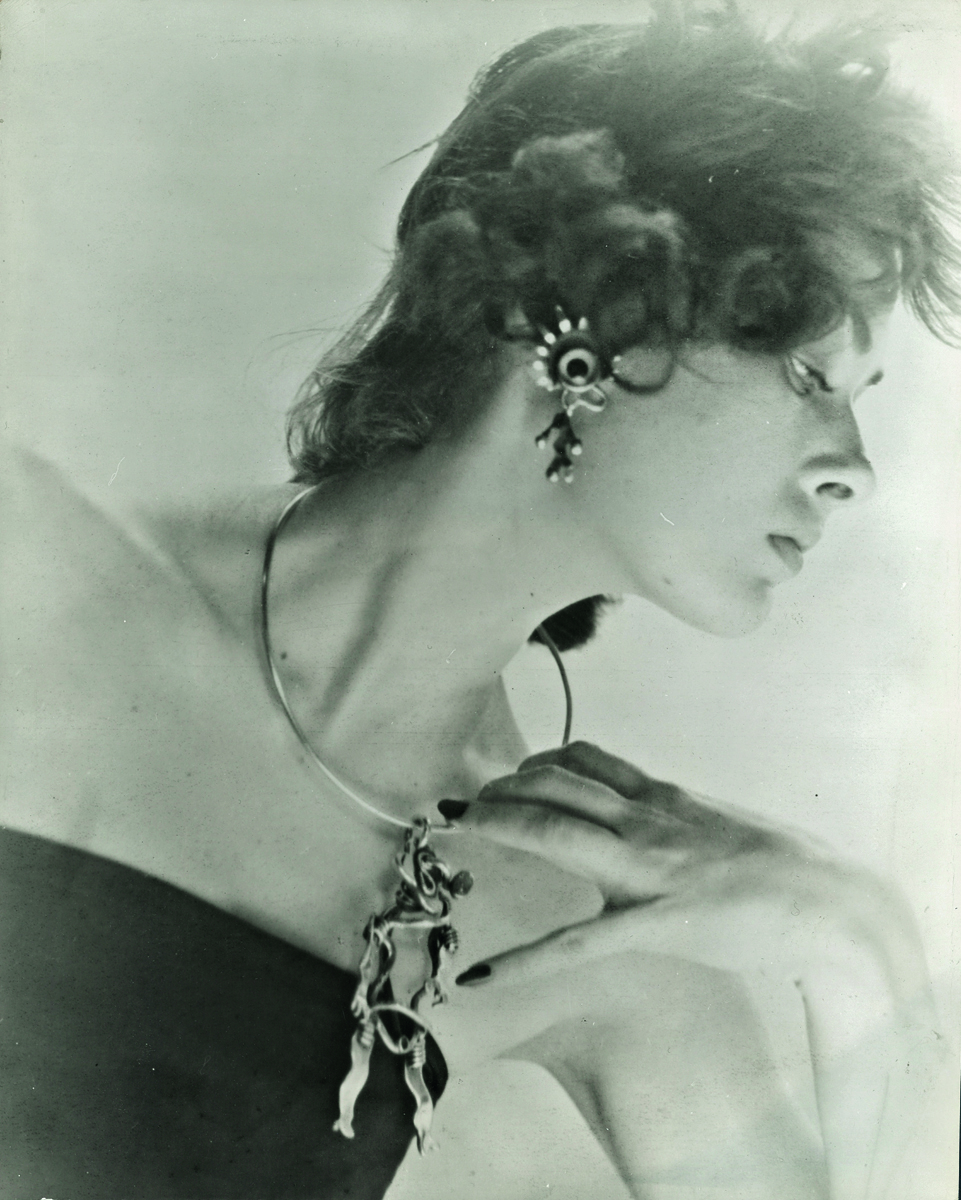 Organic shaped silver metal jeweller with coloured stones, off-white background, SAM KRAMER JEWELER ON THE EDGE in black font above.