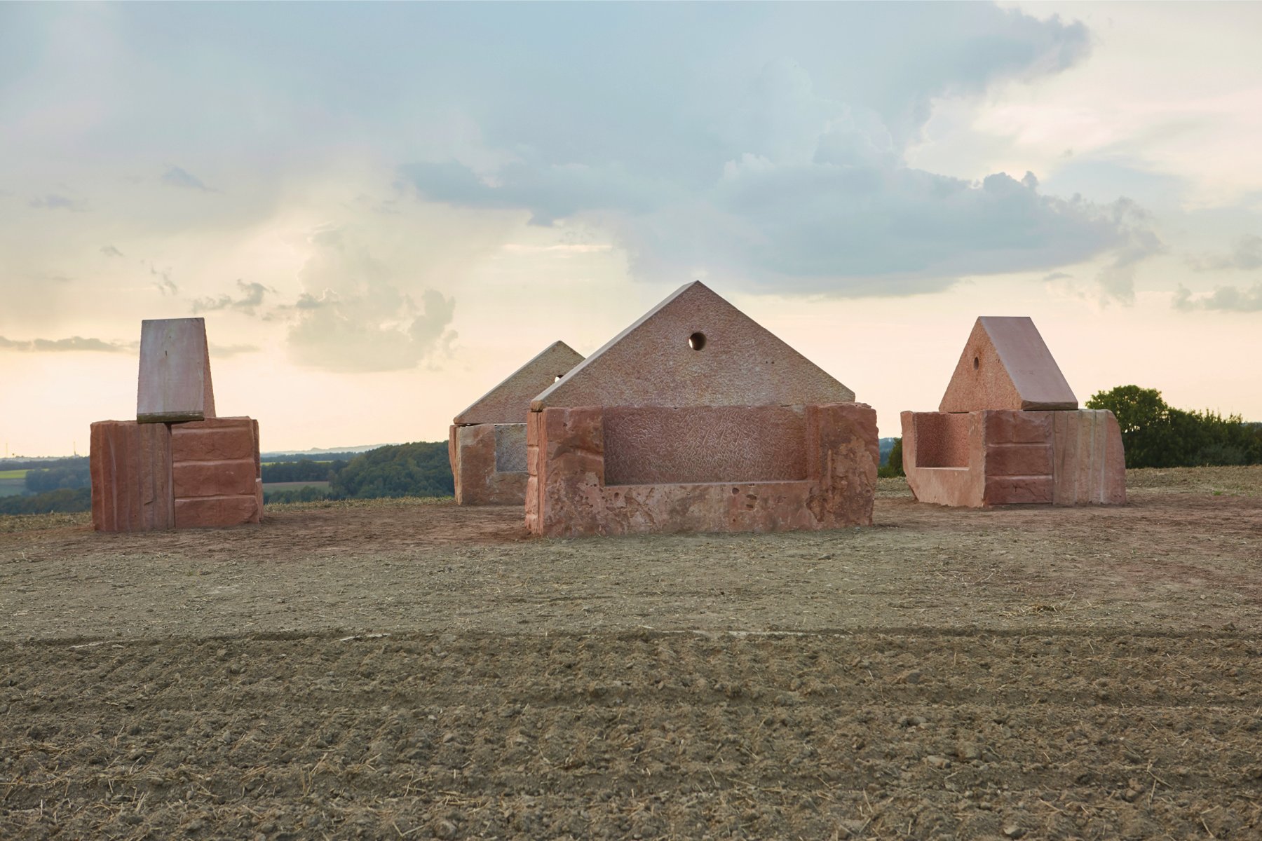 Three terracotta stone structures placed on barren land, CLAUS BURY The Poetry of Construction in white font on khaki bottom banner.