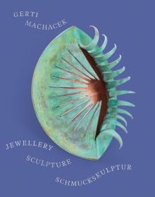 Blue book cover of Gerti Machacek, Jewellery Sculpture, featuring an oxidised copper jewellery piece, shaped like an eye, with eyelashes. Published by Arnoldsche Art Publishers.