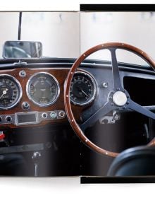 Front end of silver Aston Martin DB5, headlights on, THE ASTON MARTIN BOOK, in white font above, 'BEST SELLER' in white font to green circle.