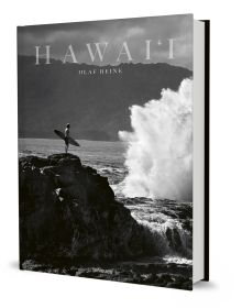 Book cover of Olaf Heine's Hawaii, with surfer standing on rocks holding surf board near the crashing waves of the sea. Published by teNeues Books.