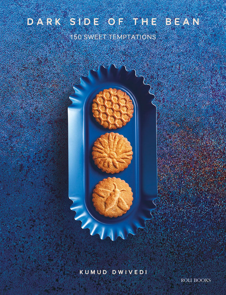 Blue baking tin with crimped ends holding 3 golden baked biscuits, on blue surface with Dark Side of the Bean 150 Sweet Temptations in white font above