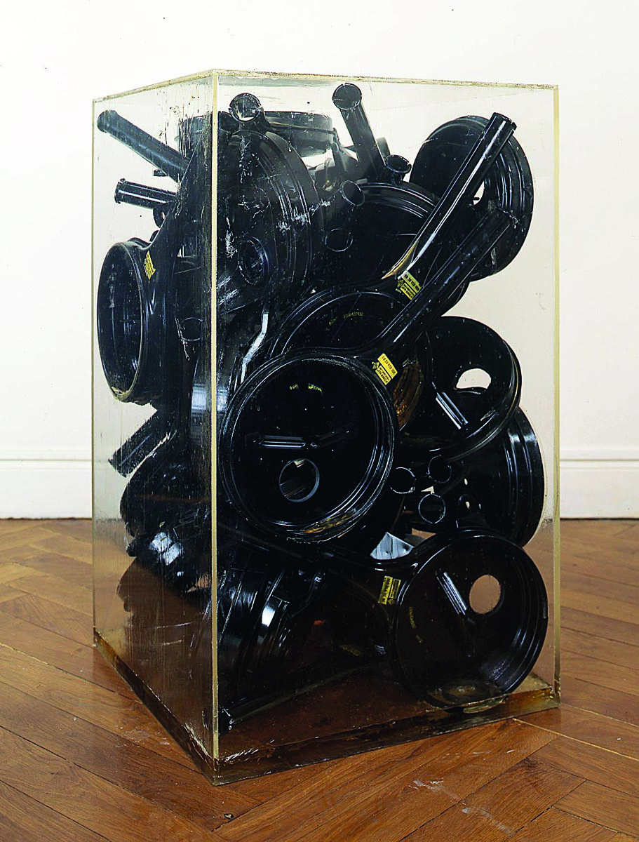 Installation sculpture of long spouted metal jugs fixed together, Germano Celant ARMAN 1955 1974 in black and blue font above.