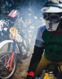 Head of biker with red helmet, peak visor and goggles, dark blue cover, Deus Ex-Machina SWANK RALLY in off white font near centre