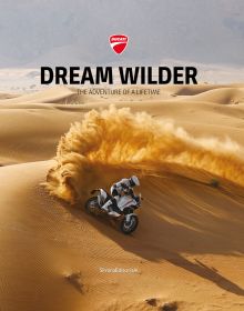 Book cover of Dream Wilder', with a Ducati DesertX off-road motorcycle with rider, speeding across a sandy desert, with sand spray behind. Published by Silvana.