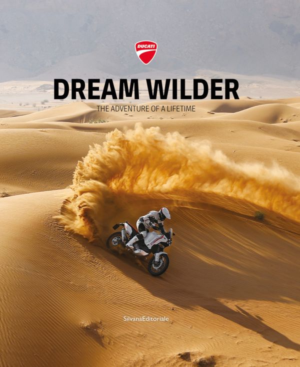 Book cover of Dream Wilder', with a Ducati DesertX off-road motorcycle with rider, speeding across a sandy desert, with sand spray behind. Published by Silvana.